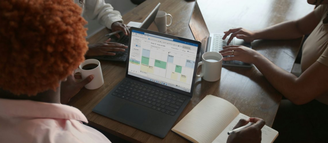 Three people in a meeting at a table discussing schedule on their Microsoft laptop